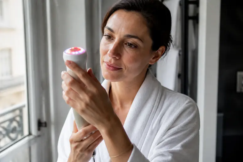 Femme utilisant un appareil de soin du visage avec différentes lumières LED dans sa salle de bain.